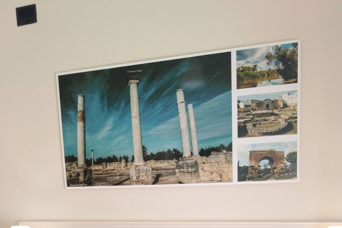 A hotel room with two beds, a large framed picture of ancient ruins, and a small table with a lamp.
