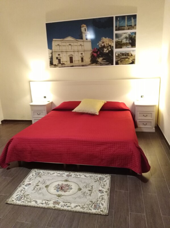 A bedroom with a red bedspread, two bedside lamps, a nightstand, a decorative rug, and a wall-mounted picture of a building.