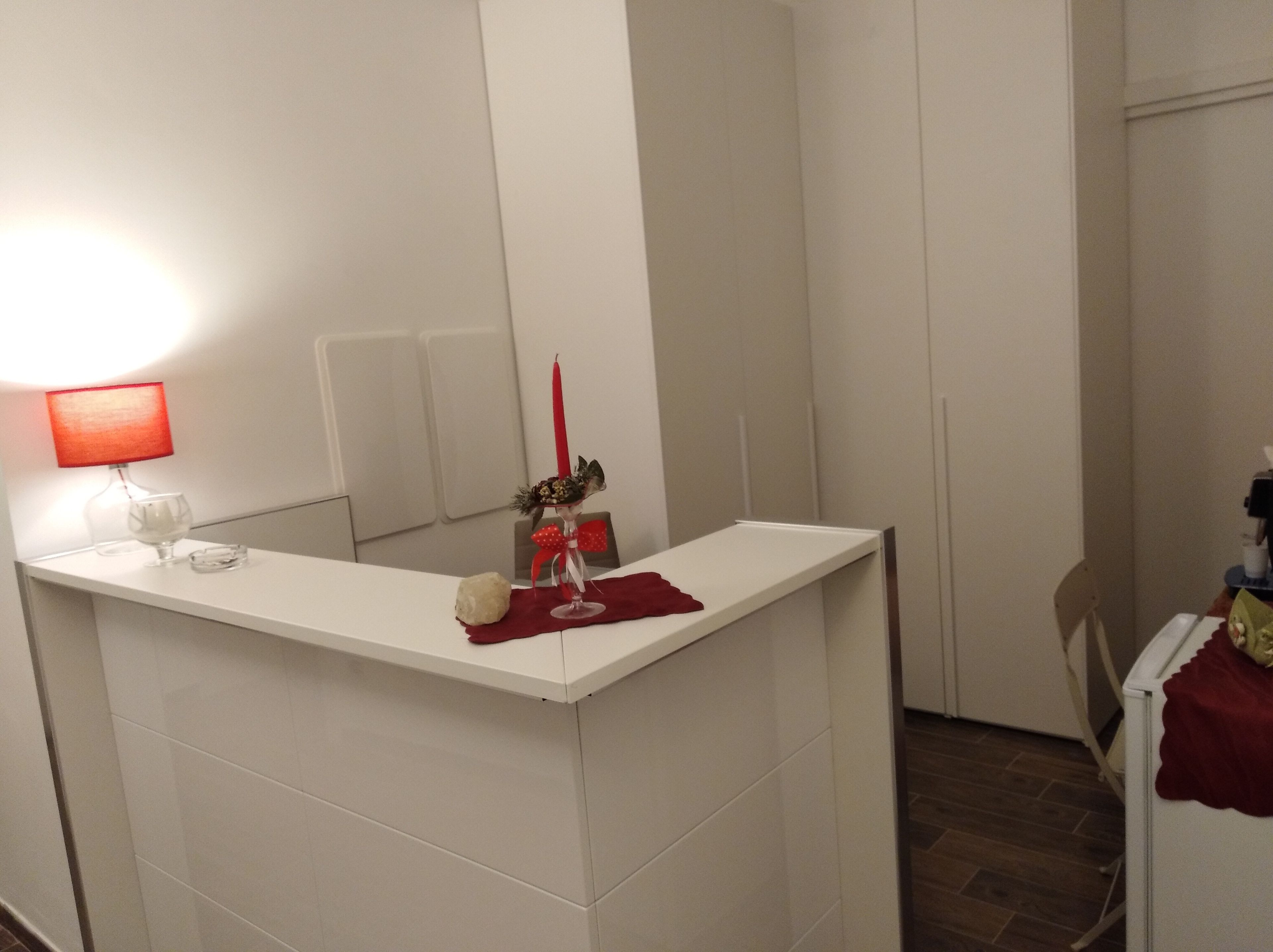 Un pequeño mostrador de recepción blanco con una lámpara roja, un florero con una vela roja y un vaso.