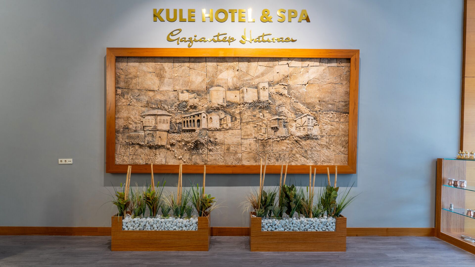 Zona d'entrada amb un cartell fixat a la paret que diu "KULE HOTEL & SPA" i un baix relleu que representa una escena històrica. Hi ha dos testos de fusta amb plantes i pedres al davant de l'escultura.