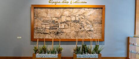 Zona d'entrada amb un cartell fixat a la paret que diu "KULE HOTEL & SPA" i un baix relleu que representa una escena històrica. Hi ha dos testos de fusta amb plantes i pedres al davant de l'escultura.