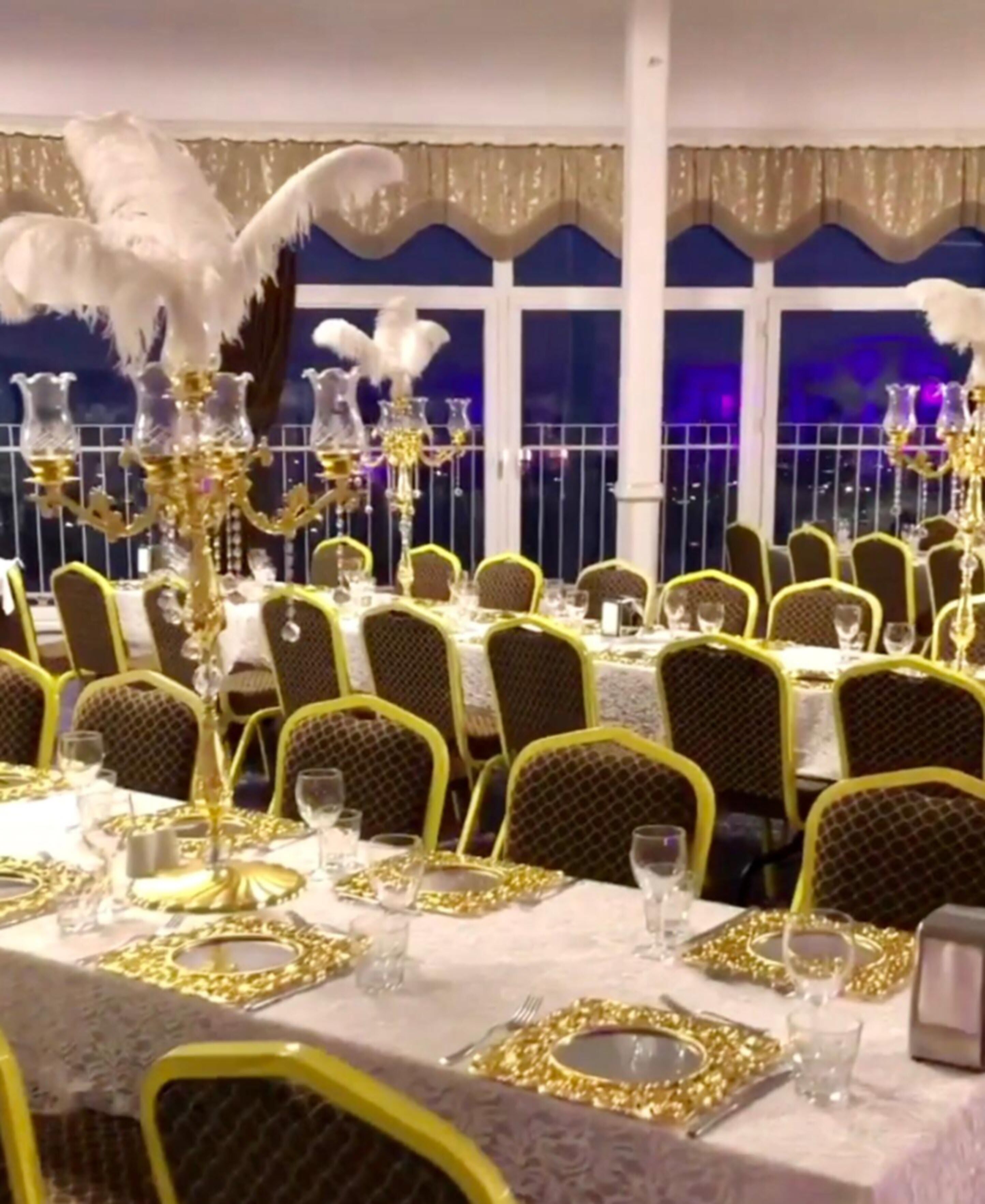 Uma sala de banquetes com mesas montadas para um evento formal, com louças douradas e decoração de penas brancas.