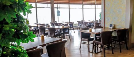 Un restaurante con mesas y sillas de madera, ventanas amplias con persianas y una planta verde en primer plano.