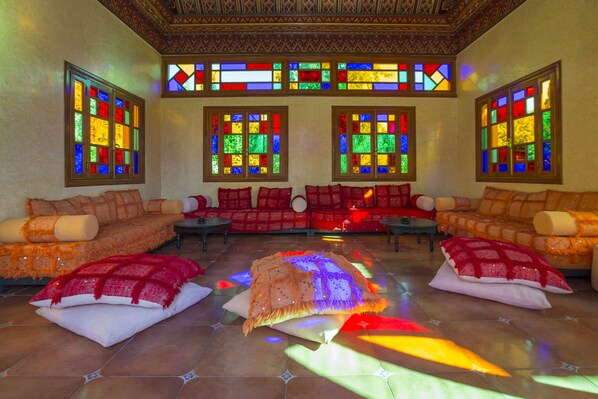 Um quarto com quatro sofás dispostos em semicírculo, cada um com um cobertor vermelho e almofadas. Há quatro janelas com vitrais coloridos, e o quarto possui piso de azulejos.