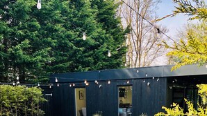 An outdoor patio area with metal chairs and tables, surrounded by greenery and a dark building structure.