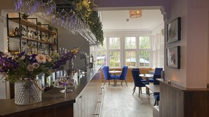 A bar area with a long counter, shelves with bottles, a vase with flowers, and blue chairs in the background.