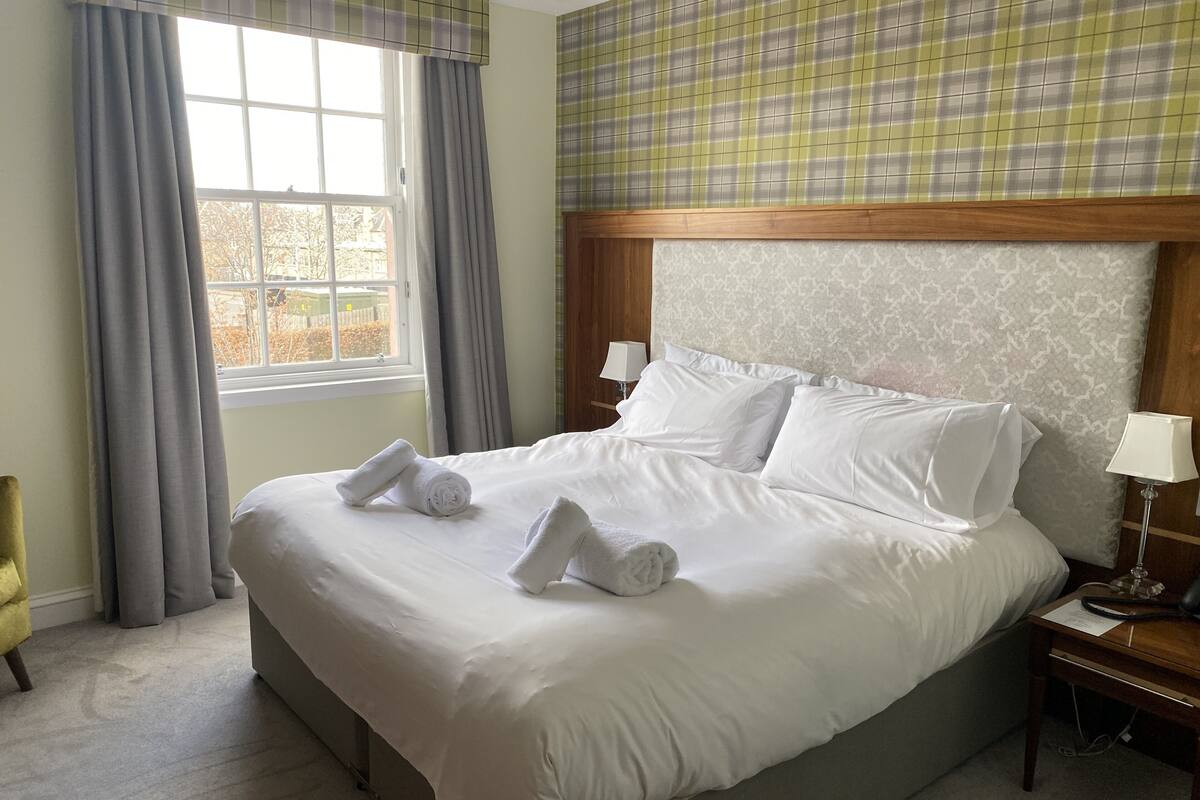 Un lit bien fait, avec du linge de lit et des serviettes blanches, une tête de lit en bois, une table de chevet avec une lampe et une fenêtre avec des rideaux.