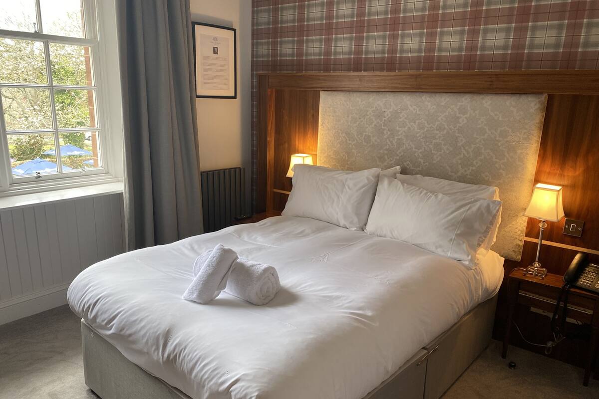 Un lit bien fait, avec du linge de lit blanc et une serviette pliée, une lampe de chevet, un certificat encadré accroché au mur et une fenêtre avec des rideaux.