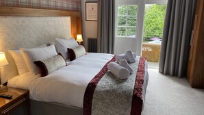 A large bed with white linens, a marble-patterned bed runner, and rolled towels on the bed. There is a nightstand with a lamp and a remote control. A framed certificate is on the wall, and a window with curtains is visible.