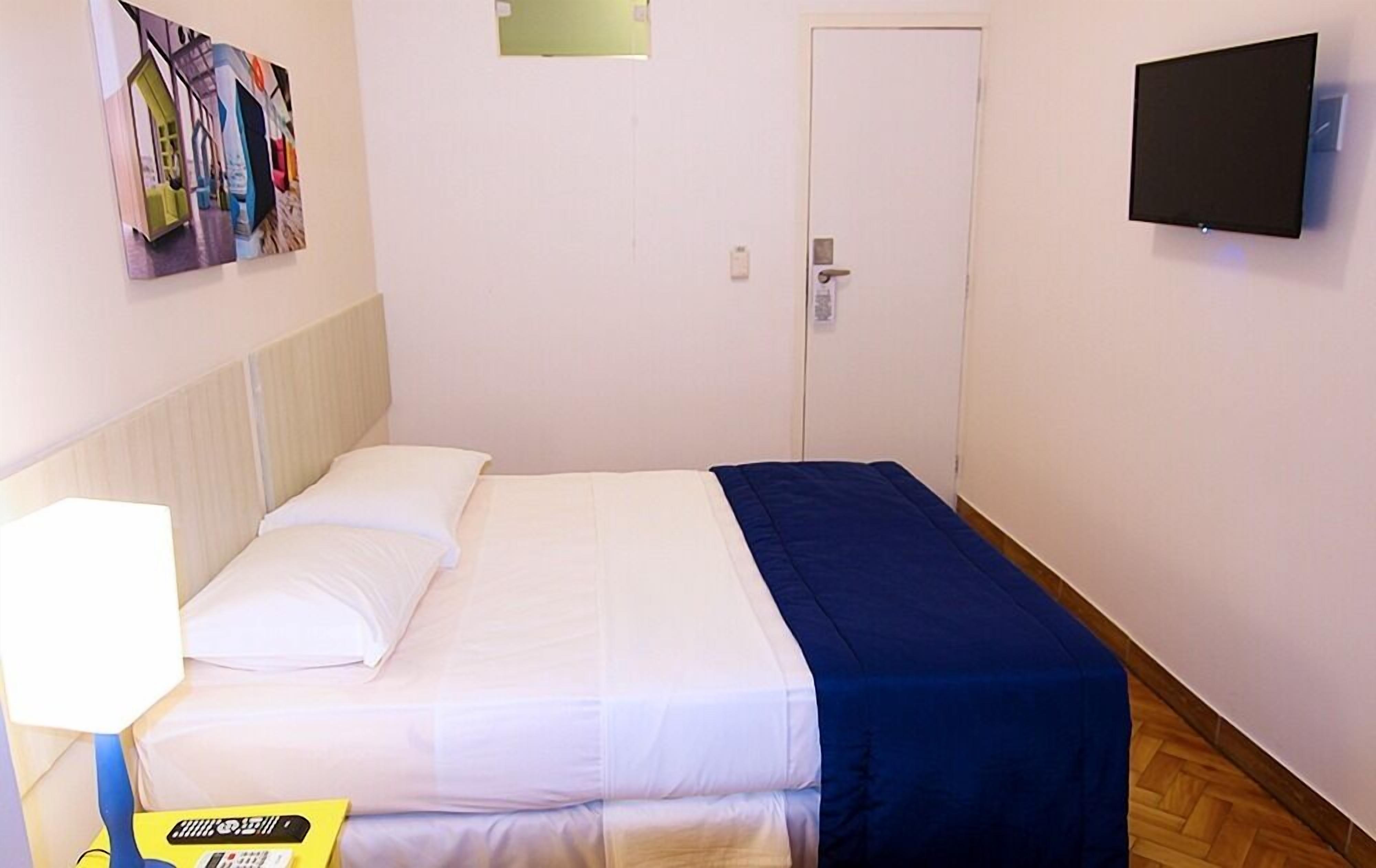 A hotel room with a bed, a bedside table with a lamp, a TV mounted on the wall, and a door.
