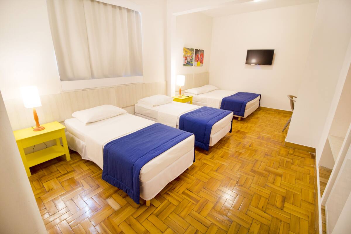 Hotellihuone, jossa on kolme sänkyä, puulattia, seinälle asennettu televisio ja ikkuna, jossa on verhot.