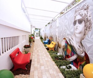 A narrow pathway with a tiled floor, surrounded by white walls and a mural of a woman. There are colorful plastic chairs and a red chair along the path. - Hotel Villa Smart (Fortaleza)