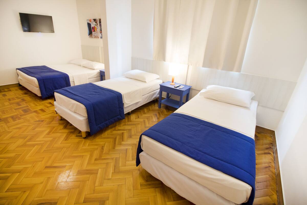 Hotellihuone, jossa on kolme erillistä sänkyä, puulattia, pieni pöytä lampun kanssa ja seinälle asennettu taulutelevisio.