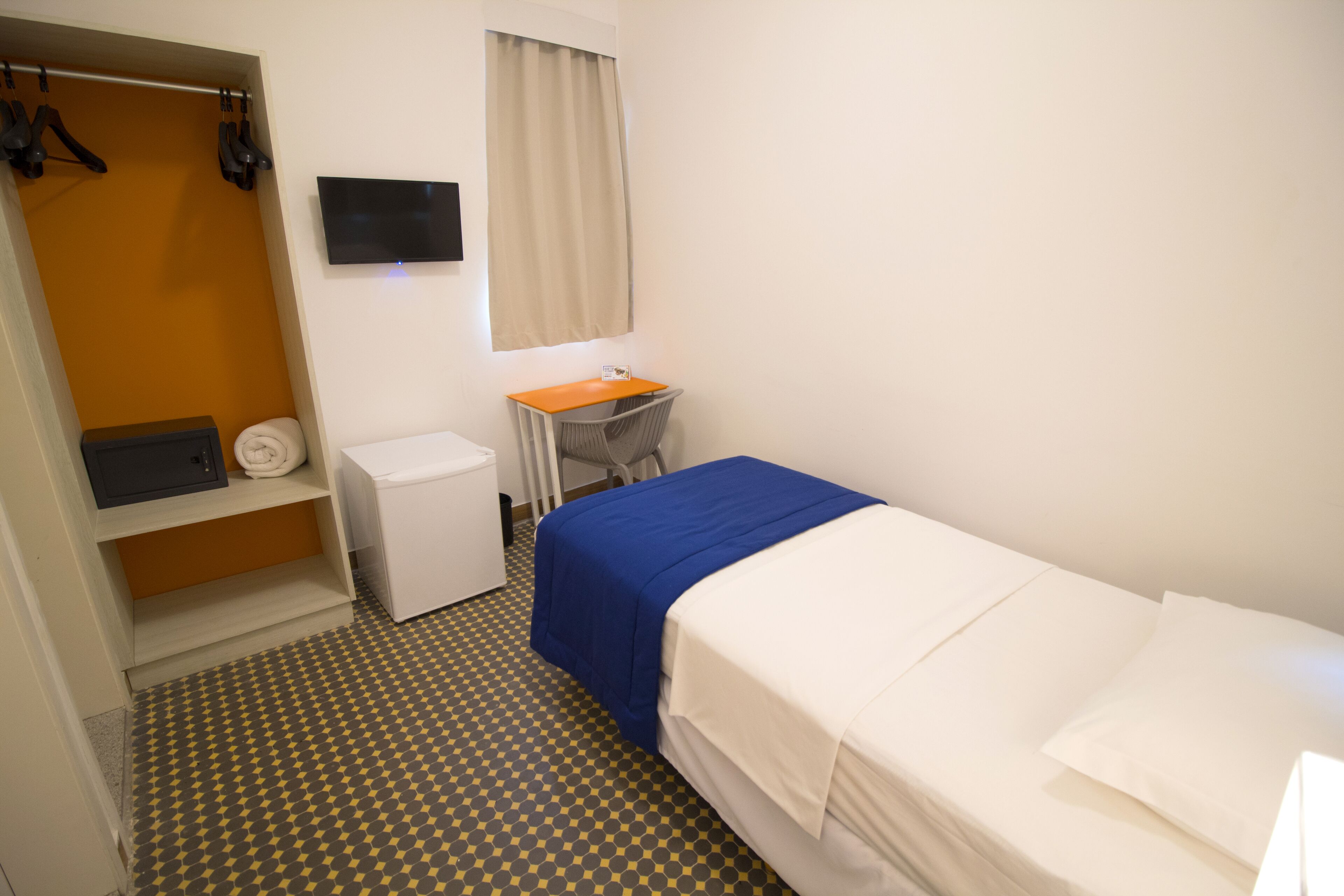 A hotel room with a bed, a desk, a chair, a small table, a TV, and a wardrobe.