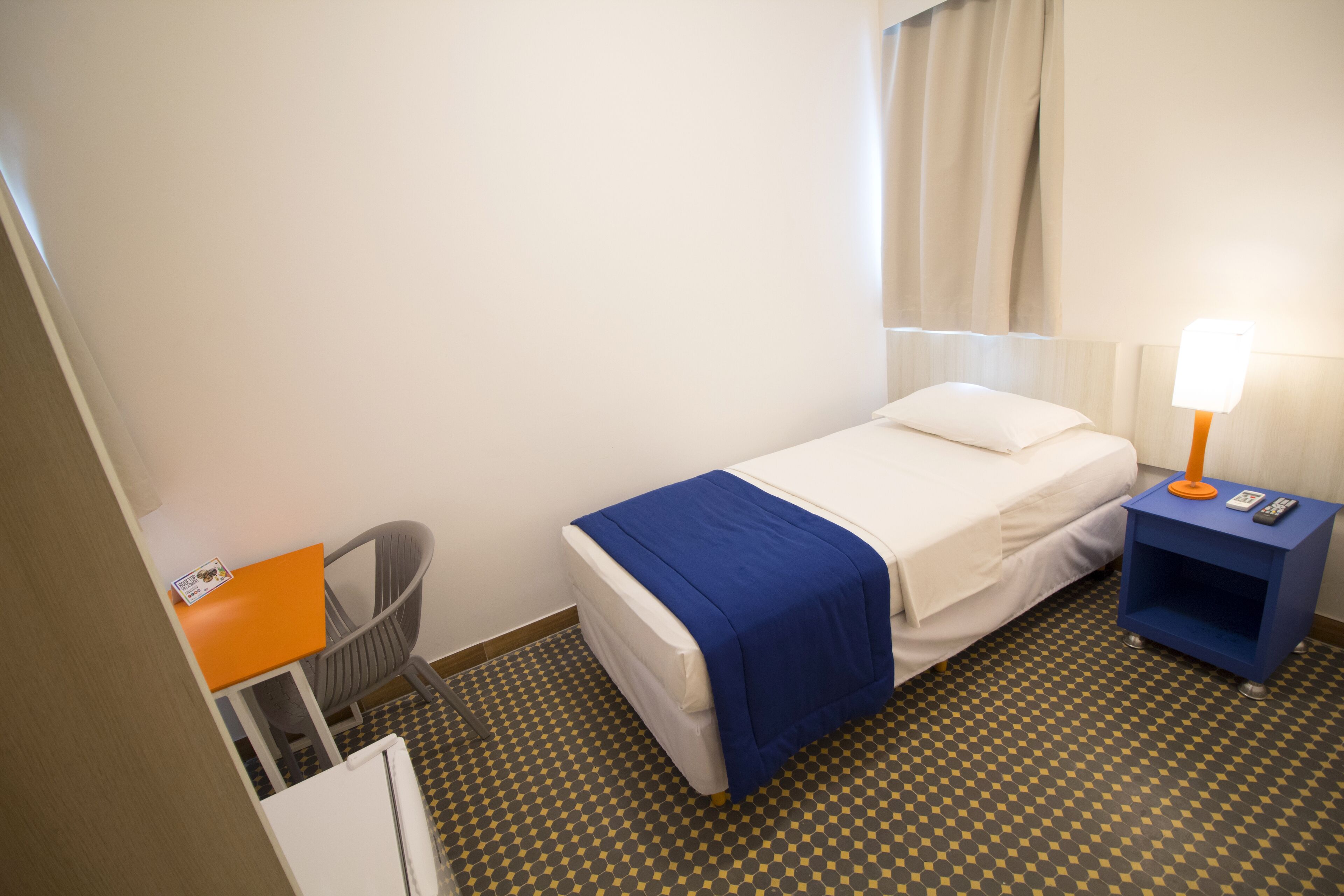A hotel room with a bed, a desk, a chair, a lamp, and a small table.