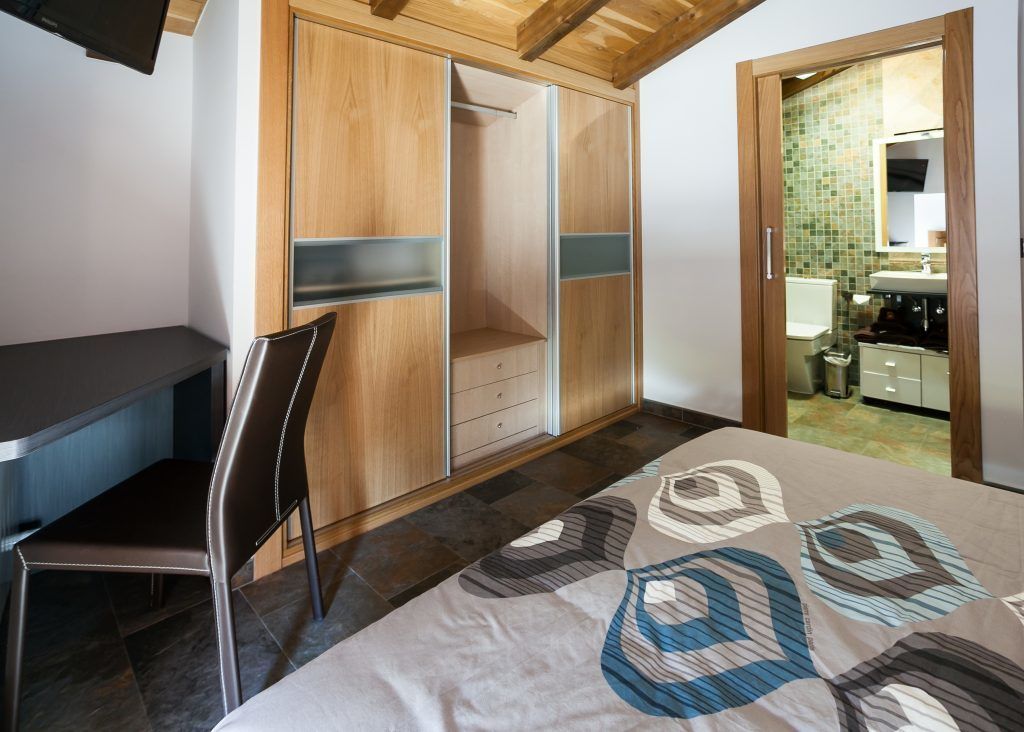 Quarto com cama, escrivaninha, cadeira, guarda-roupa e banheiro visível por uma porta aberta.