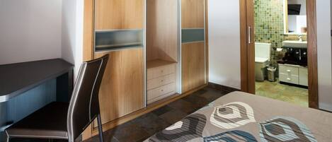 Quarto com cama, escrivaninha, cadeira, guarda-roupa e banheiro visível por uma porta aberta.