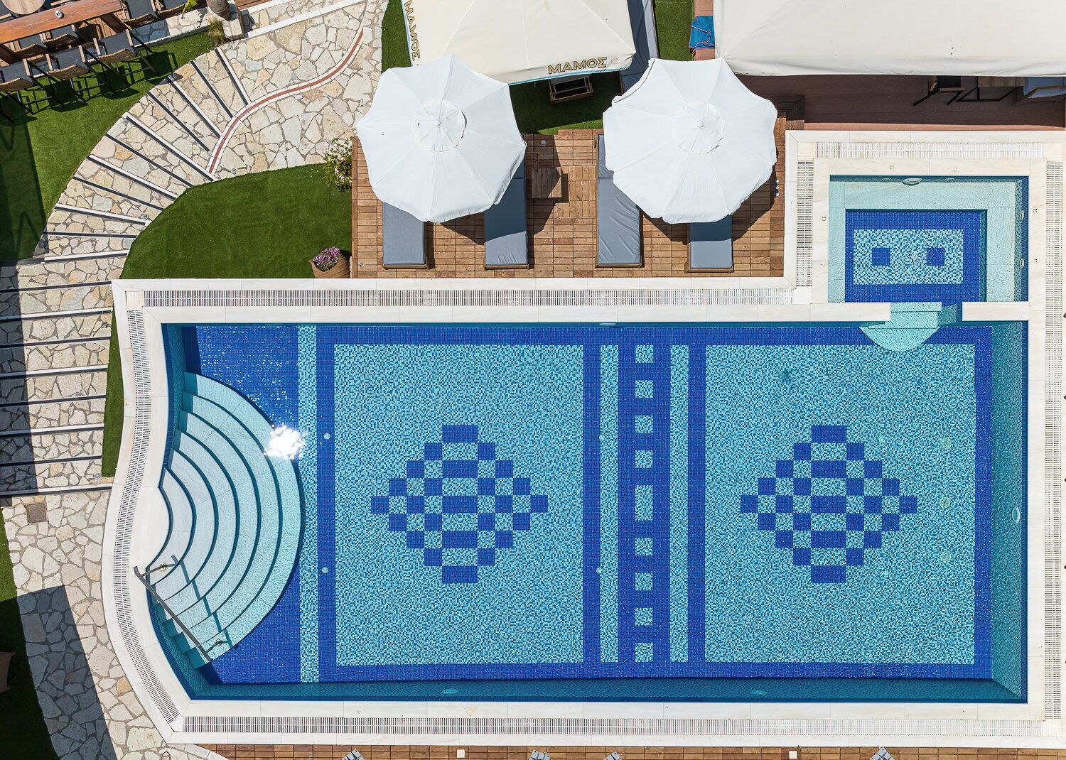 A large outdoor swimming pool with a geometric tile pattern, surrounded by a stone deck and two white umbrellas.