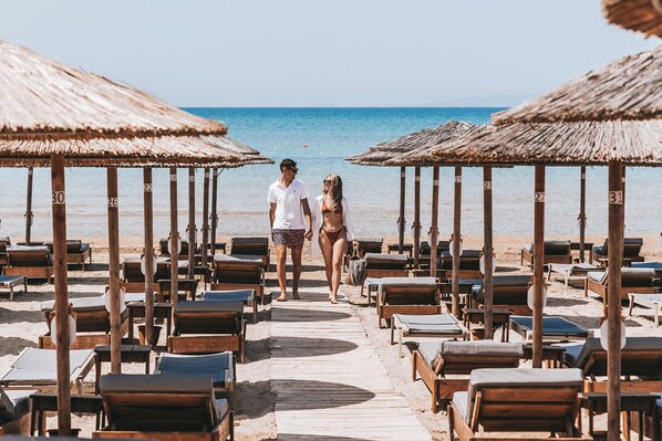 A beach with wooden lounge chairs and thatched umbrellas, arranged in rows along a wooden walkway. - Hotel Almira (Pineios)