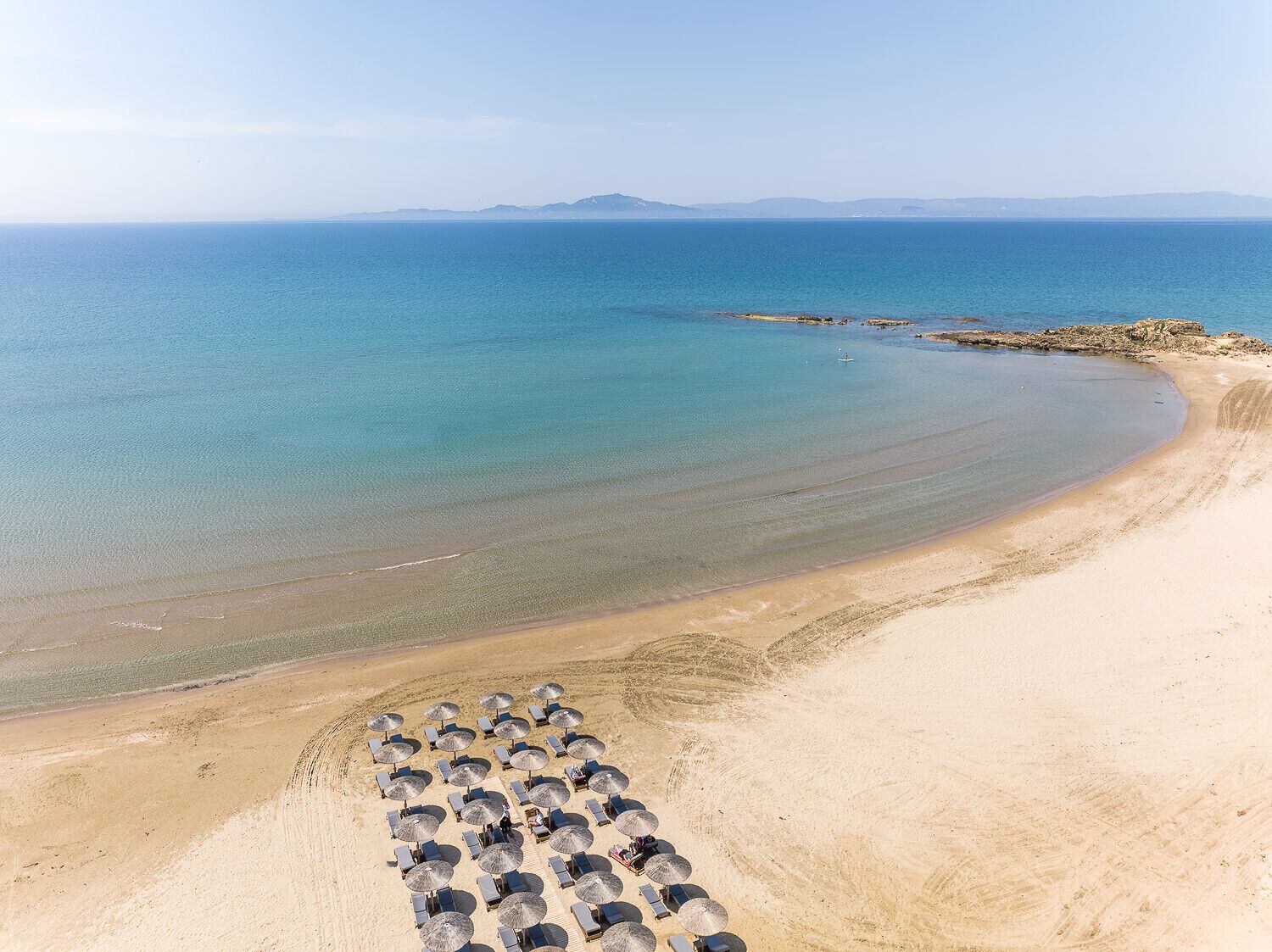Una playa con filas de tumbonas y sombrillas, un mar azul cristalino y montañas a lo lejos.