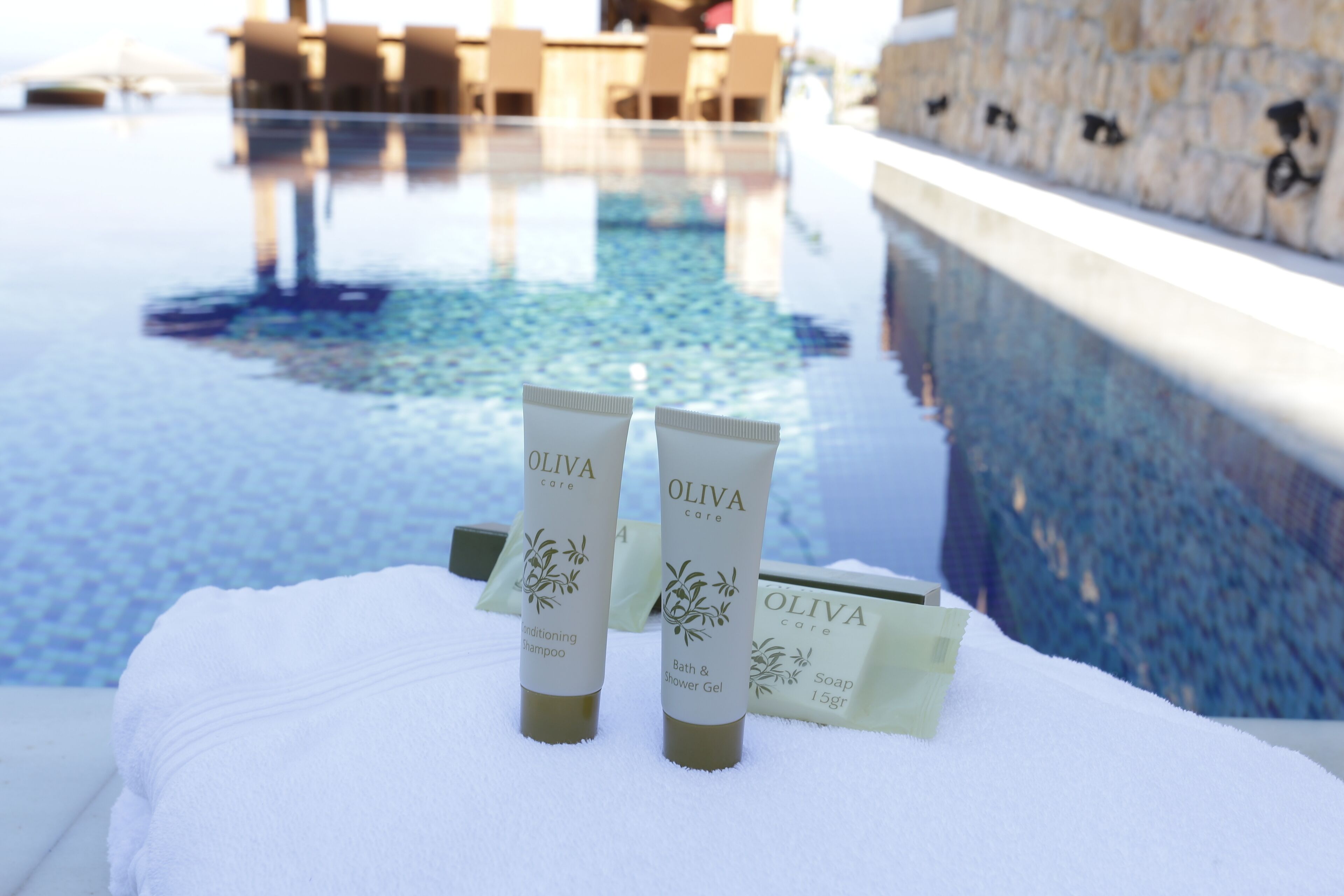 Zona de piscina con dos botellas de productos OLIVA sobre una toalla blanca.