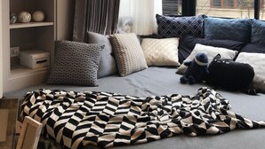 A modern bedroom with a large window, a sofa with multiple cushions, a bedside table, and a wall-mounted shelf with decorative items. - Stay in Style Family Room at Nimman R201 (Chiang Mai)