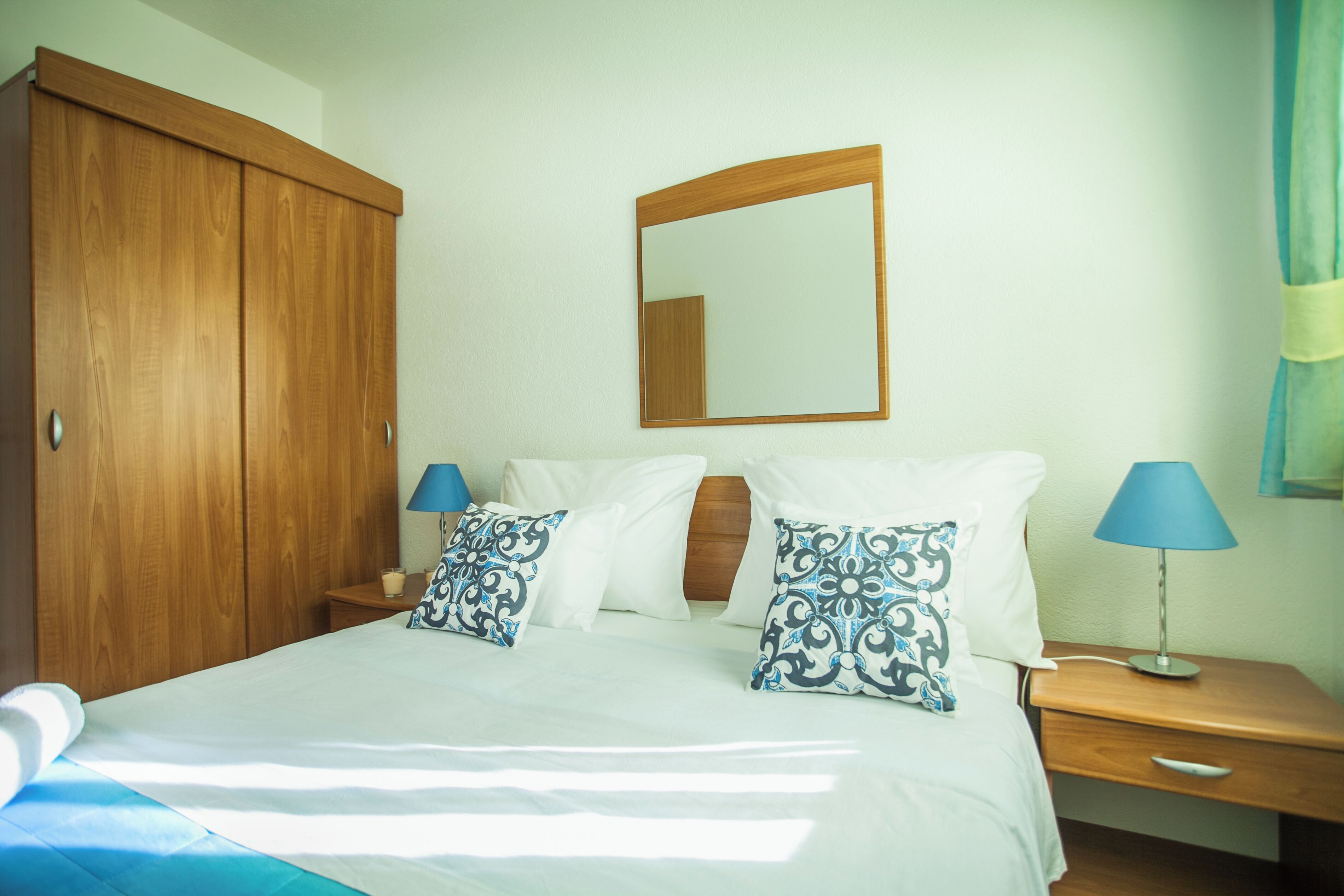 A bedroom with a wooden wardrobe, a bed with white bedding and patterned pillows, a nightstand with a lamp, and a mirror on the wall.