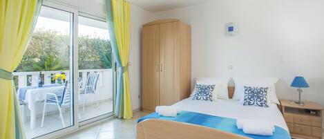 A bedroom with a bed, a wooden wardrobe, a nightstand with a lamp, and a balcony with a table and chairs.