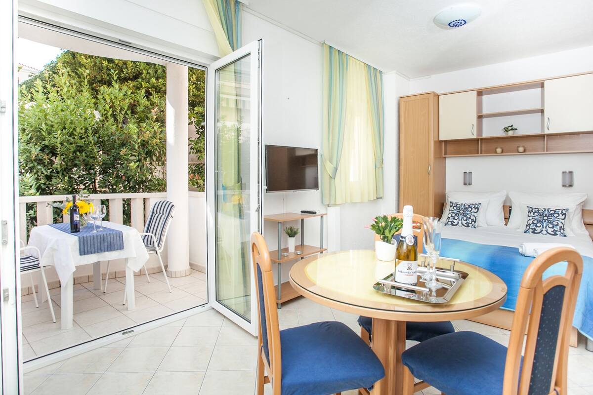 A hotel room with a balcony, a dining area, a bed with a TV, and a small table with a bottle and glasses.