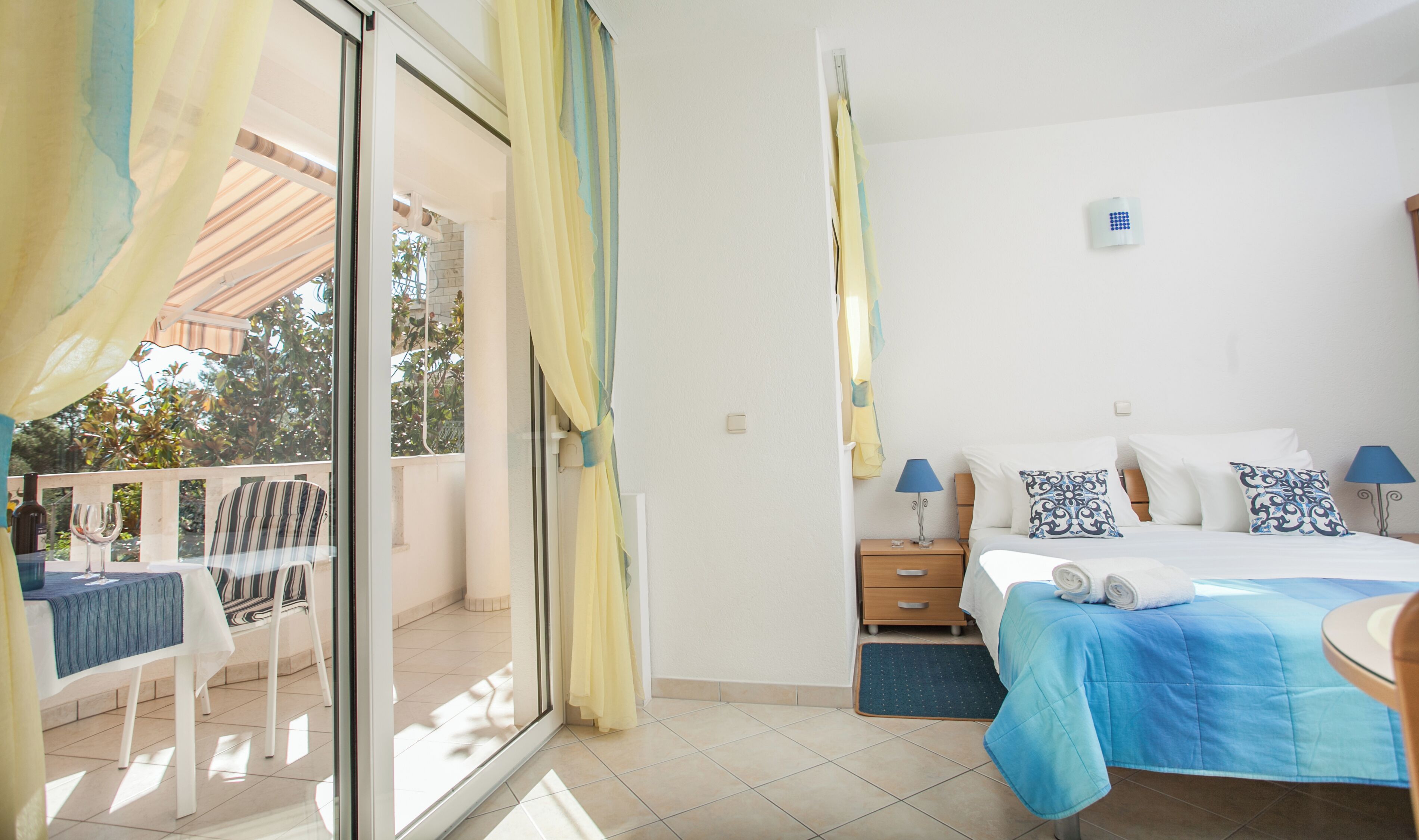 A hotel room with a bed, a small table with a chair, a balcony with curtains, and a view of trees outside.