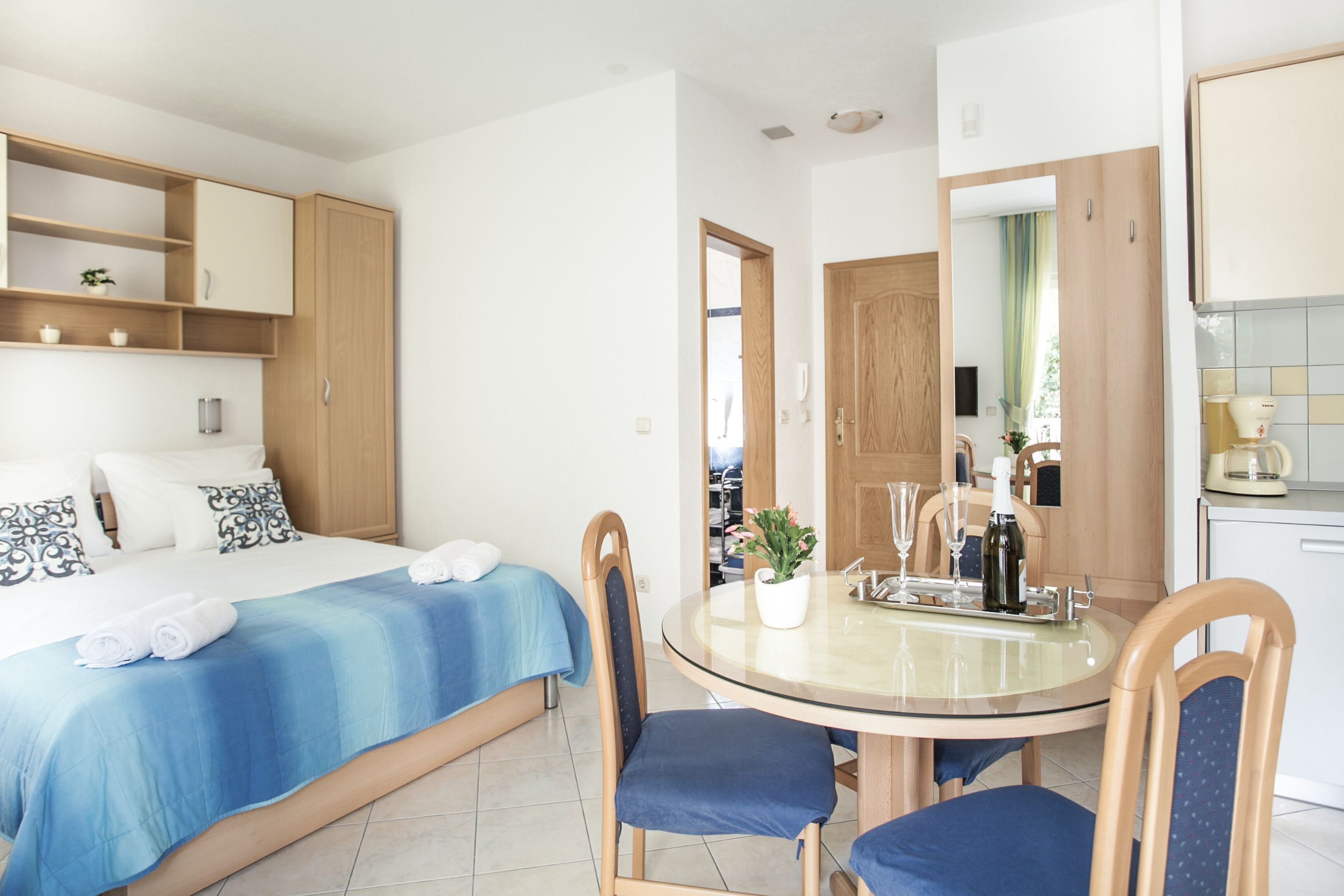A hotel room with a bed, a round dining table with chairs, a small kitchen area, and a bathroom visible through an open door.