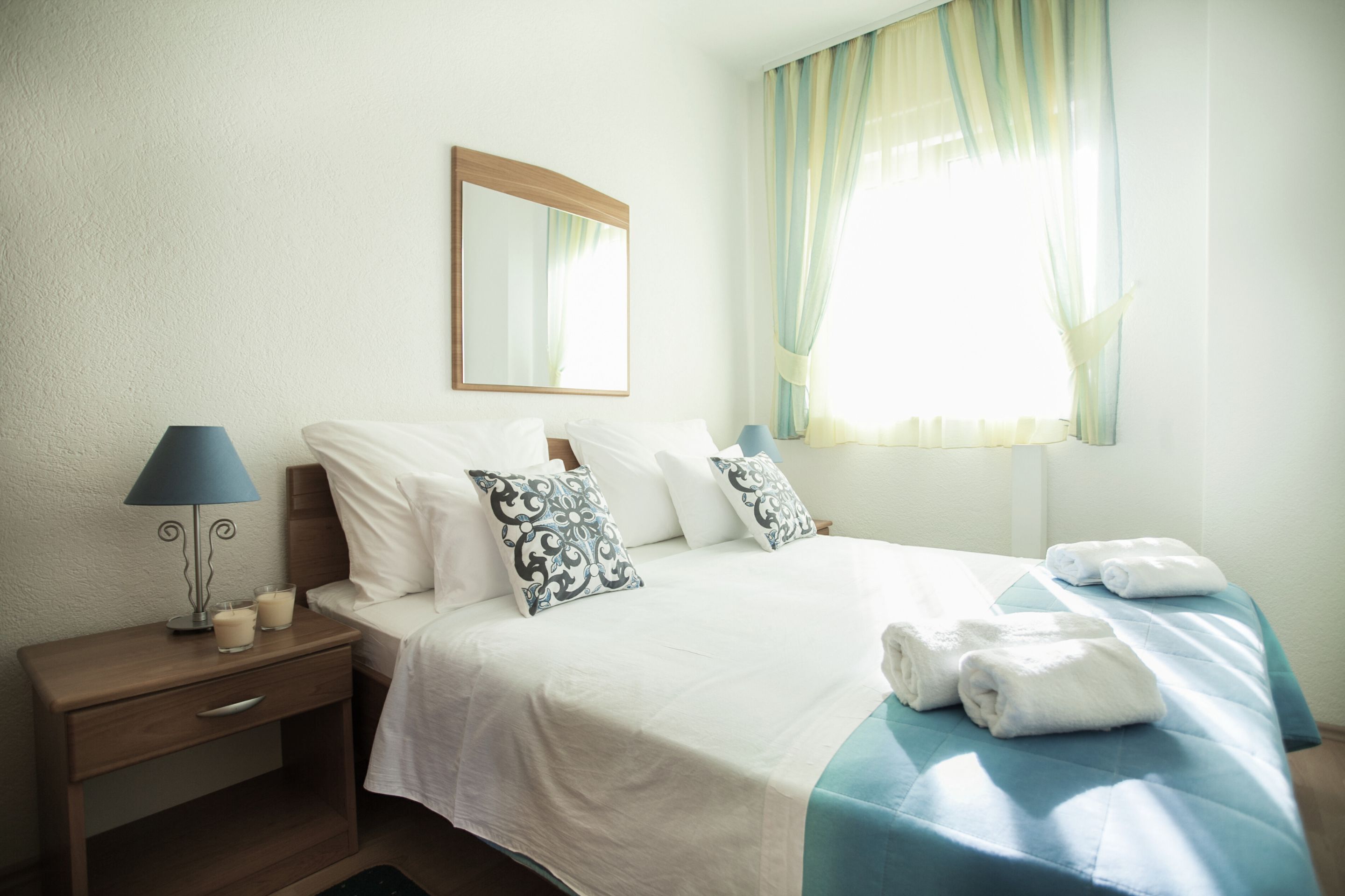 A neatly made bed with white and blue bedding, a bedside lamp, and a window with curtains.