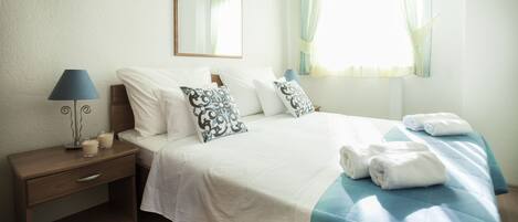 A neatly made bed with white and blue bedding, a bedside lamp, and a window with curtains.
