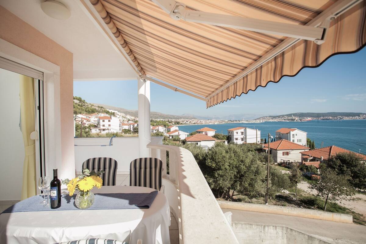 A balcony with a table set for two, a bottle of wine, and a vase with flowers. The balcony overlooks a coastal town with houses and the sea.