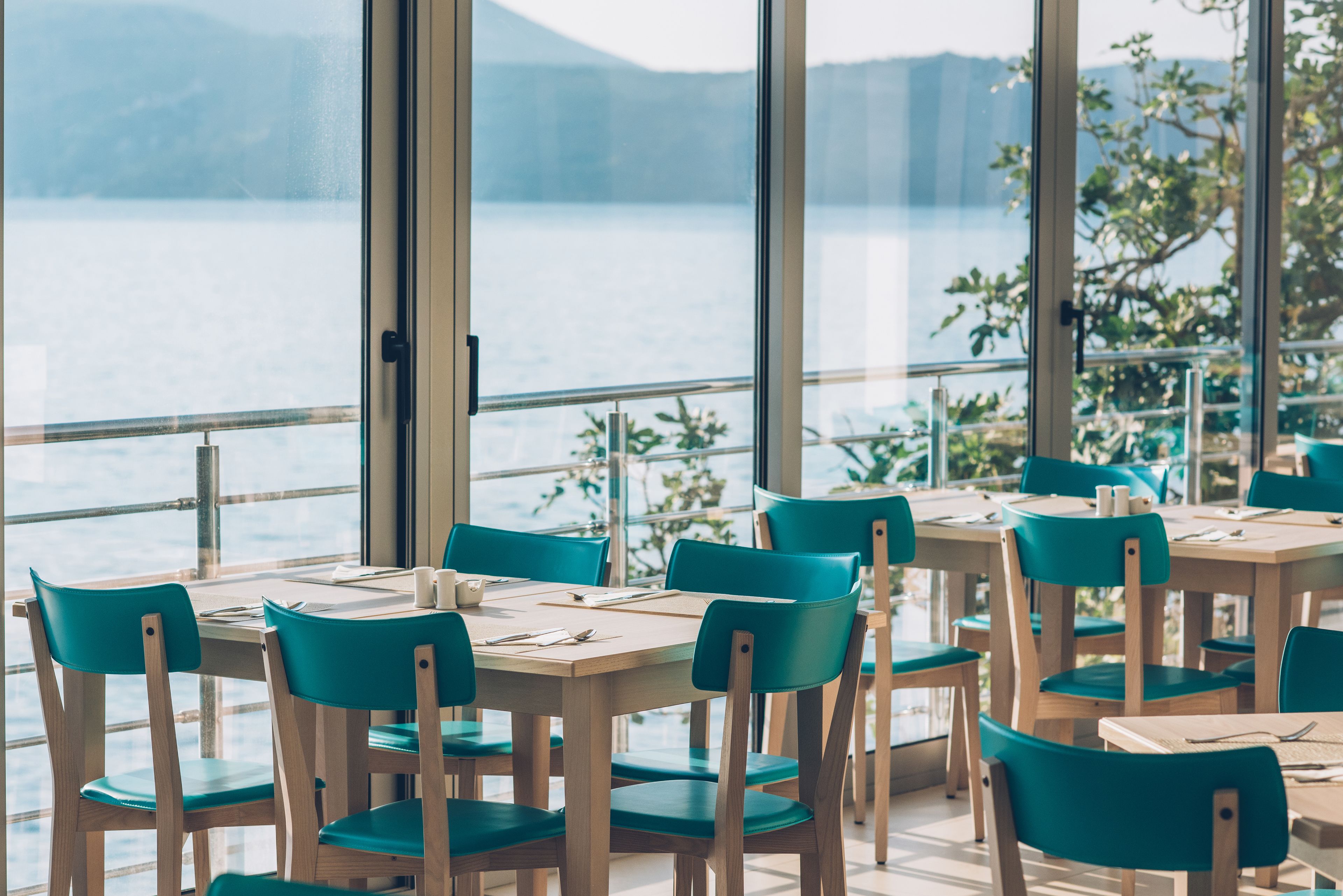 A restaurant with wooden tables and teal chairs, overlooking a body of water.