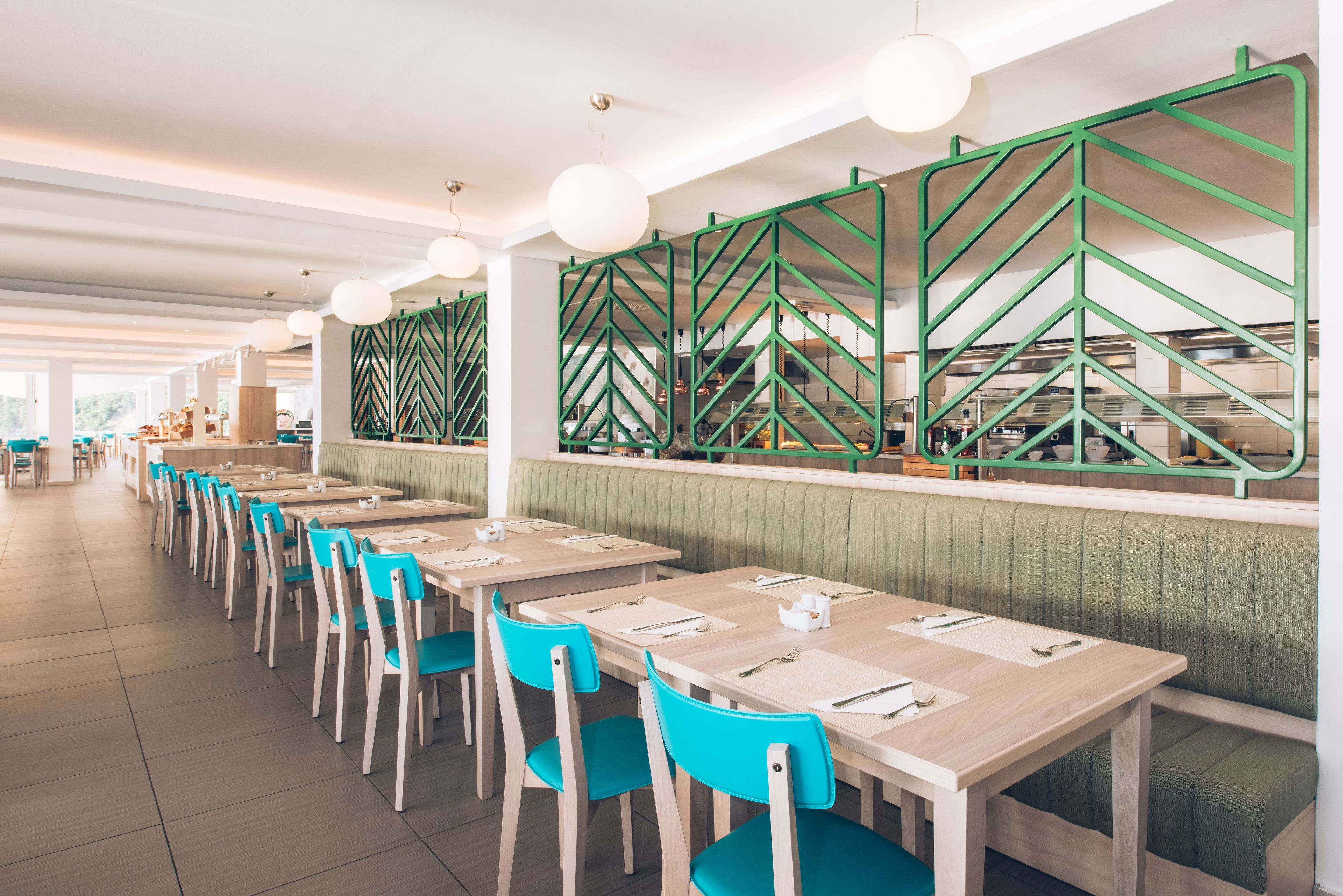 A modern restaurant with long wooden tables and teal chairs, green chevron-patterned partitions, and pendant lighting.
