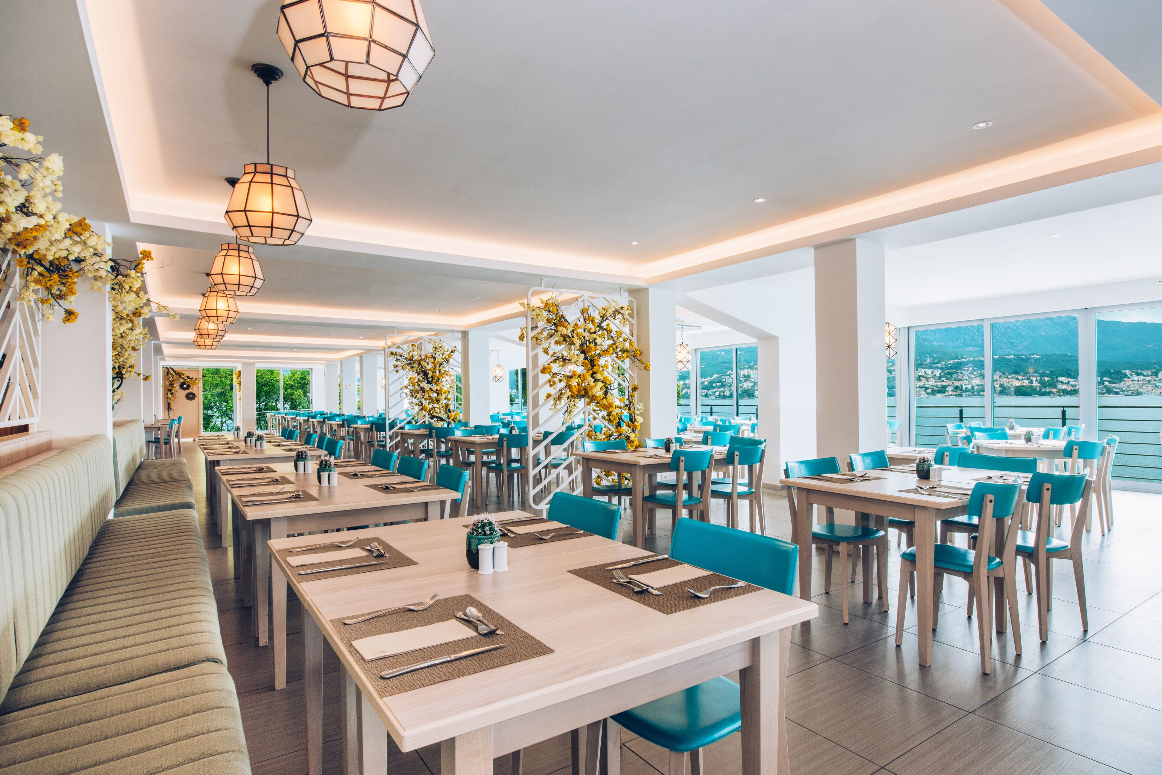 Ein modernes Restaurant mit langen Gemeinschaftstischen und Sitzgelegenheiten, große Fenster mit Blick auf die Stadt und dekorative Blumenarrangements.