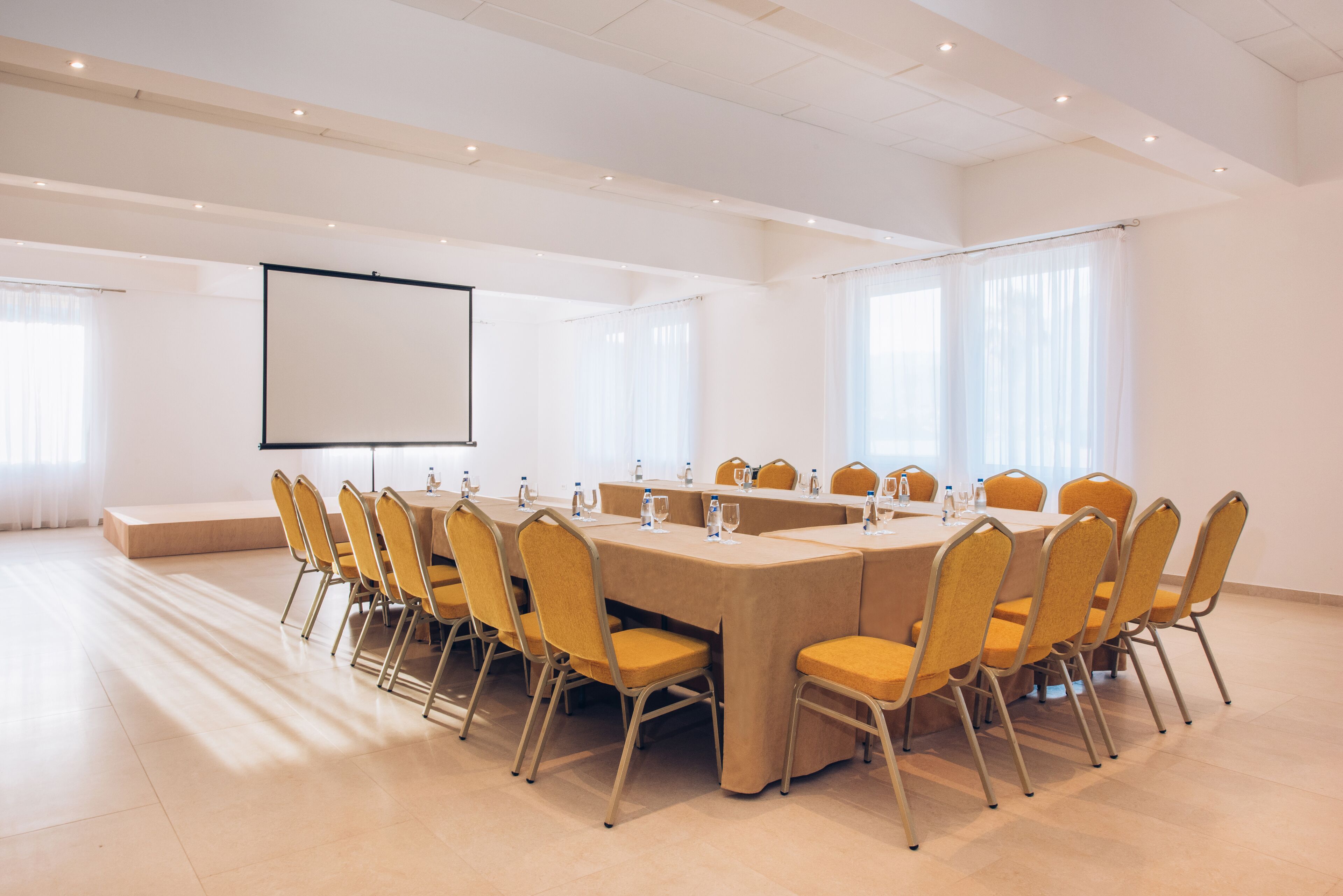 Een vergaderzaal met een lange tafel, gele stoelen, een projectiescherm en grote ramen.