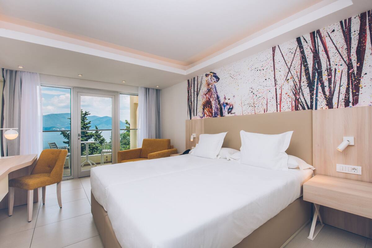 Quarto de hotel com uma cama grande, uma escrivaninha, uma cadeira e vista para as montanhas pela varanda.