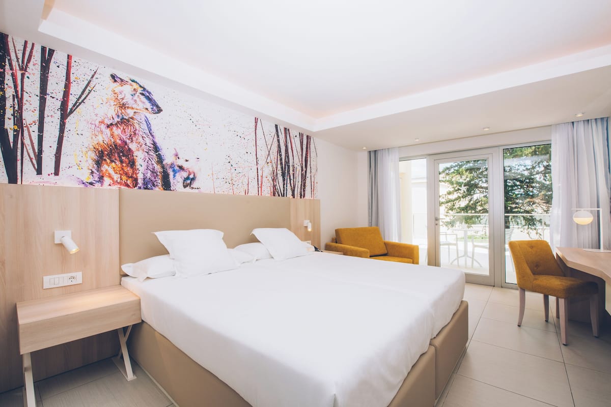Quarto de hotel moderno com uma cama grande, uma escrivaninha, uma cadeira e uma varanda com vista para árvores.