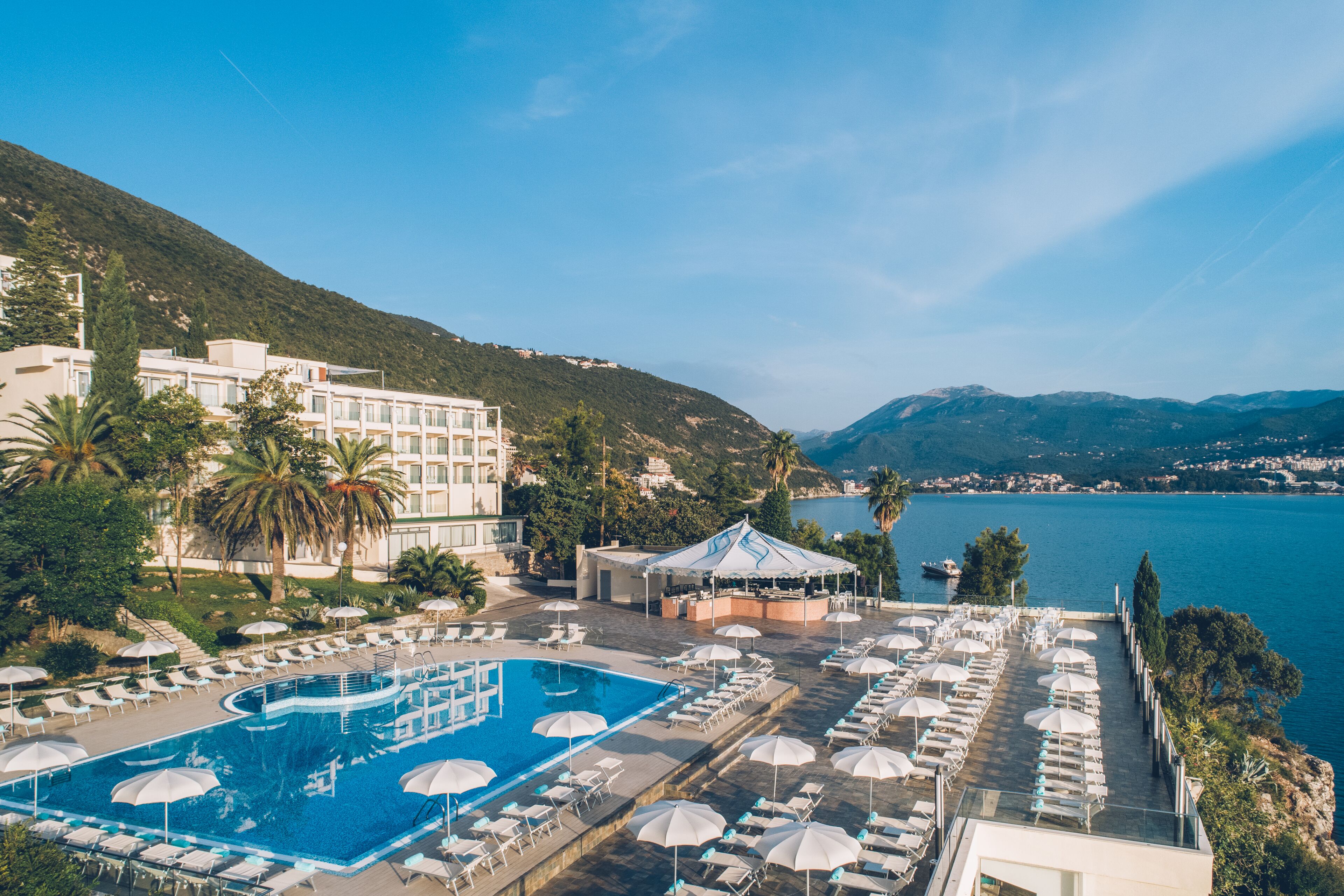 Ein Resort mit Swimmingpool, Liegestühlen, Sonnenschirmen und Blick auf Meer und Berge.