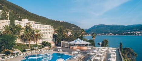 Ein Resort mit Swimmingpool, Liegestühlen, Sonnenschirmen und Blick auf Meer und Berge.