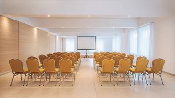 Un salón de conferencias con filas de sillas orientadas hacia una pantalla de proyección, ventanas amplias y un revestimiento de paneles de madera.