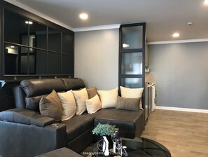 A modern living room with a dark leather sofa, multiple cushions, a glass coffee table with a vase and wine glasses, and a large window with blinds.