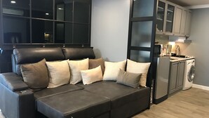 A modern living room with a sectional sofa, a built-in window, and a compact kitchen area with a washing machine. - Stay in Style The Heart of Nimman R306A (Chiang Mai)
