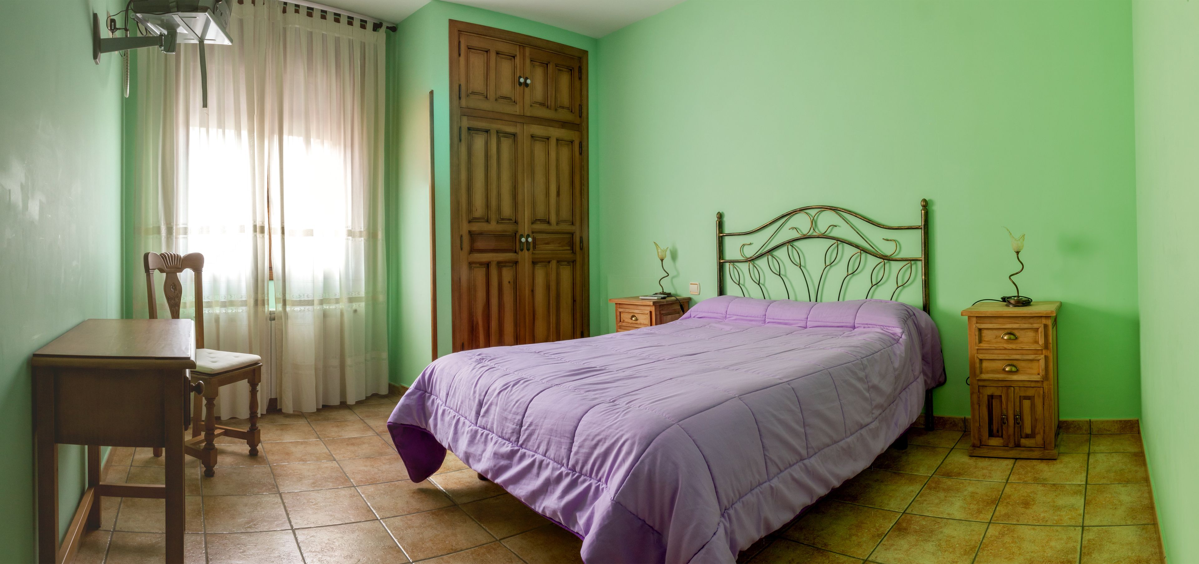 A bedroom with a bed, a chair, a nightstand, a wooden wardrobe, and a window with curtains.