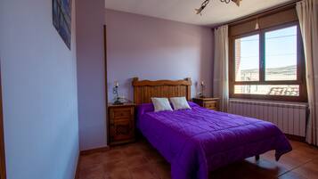 Een slaapkamer met een houten hoofdbord, een paars dekbed, twee nachtkastjes met lampen, een raam met gordijnen en uitzicht op buiten.