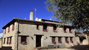 A two-story stone building with red shutters, a visible chimney, and a small garden area with a bench and potted plants. - Hotel Rural Tirontillana (Cuellar)