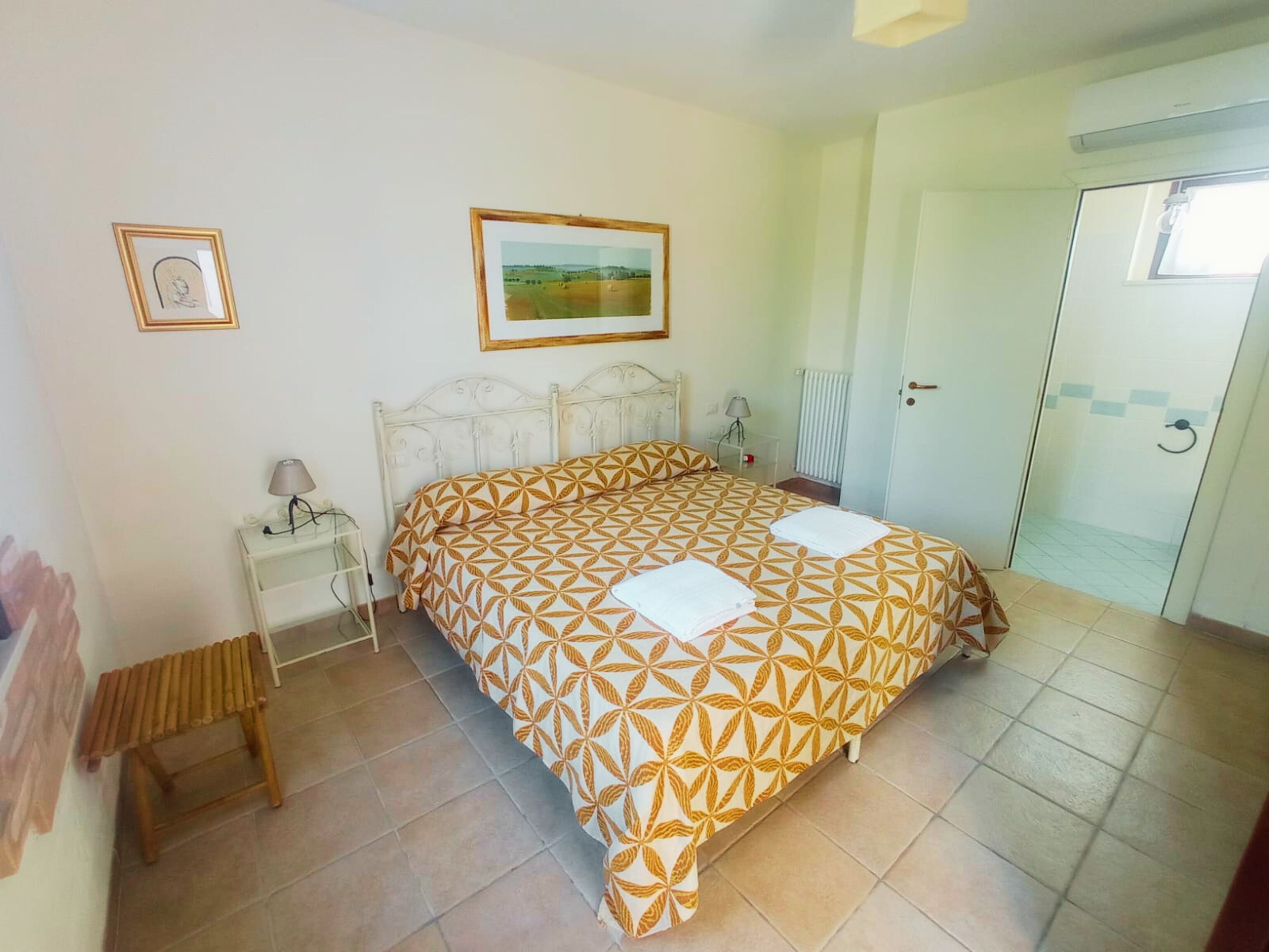 A bedroom with a bed, a nightstand, a lamp, a framed picture, and a door leading to a bathroom.