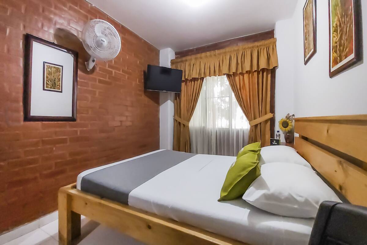 A room with a wooden bed, a wall-mounted TV, a standing fan, and a window with curtains.